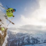 Ein Skifahrer in leuchtender Ausrüstung springt von einer schneebedeckten Klippe, im Hintergrund sind Berge und ein bewölkter Himmel zu sehen.