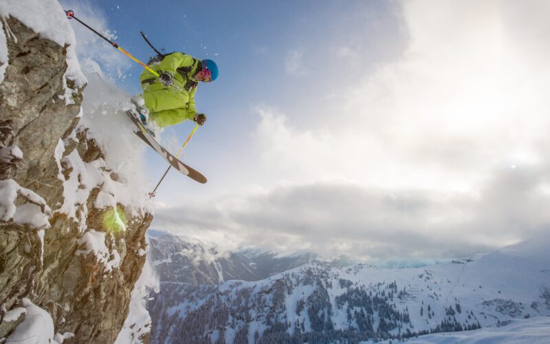 Ein Skifahrer in leuchtender Ausrüstung springt von einer schneebedeckten Klippe, im Hintergrund sind Berge und ein bewölkter Himmel zu sehen.
