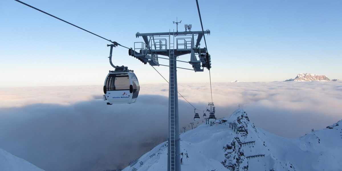Eine Seilbahn fährt über schneebedeckte Berge und ein wolkenverhangenes Tal, im Vordergrund steht ein hoher Stützturm.