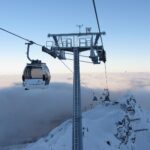 Eine Seilbahn fährt über schneebedeckte Berge und ein wolkenverhangenes Tal, im Vordergrund steht ein hoher Stützturm.