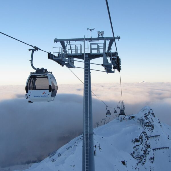 Eine Seilbahn fährt über schneebedeckte Berge und ein wolkenverhangenes Tal, im Vordergrund steht ein hoher Stützturm.