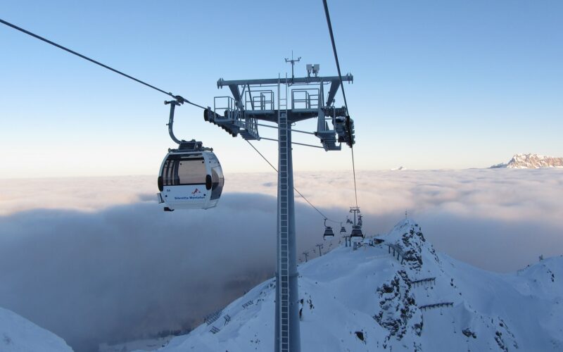 Eine Seilbahn fährt über schneebedeckte Berge und ein wolkenverhangenes Tal, im Vordergrund steht ein hoher Stützturm.