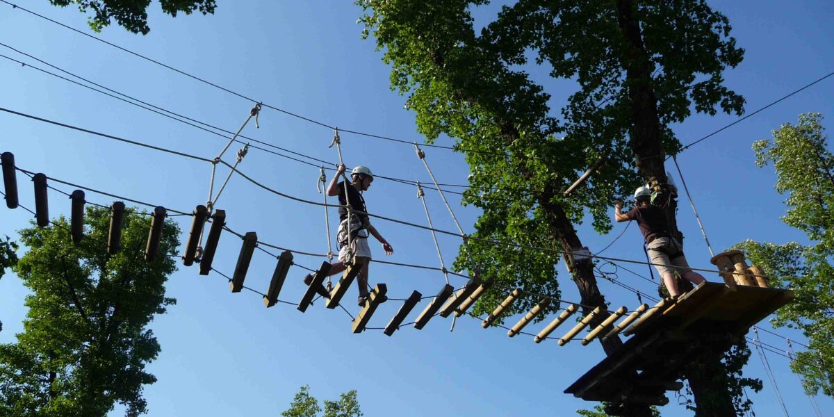 Zwei Personen mit Helmen überqueren an einem klaren, sonnigen Tag eine Seilbrücke hoch zwischen den Bäumen in einem Baumkronen-Abenteuerpark.