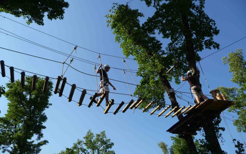Zwei Personen mit Helmen überqueren an einem klaren, sonnigen Tag eine Seilbrücke hoch zwischen den Bäumen in einem Baumkronen-Abenteuerpark.