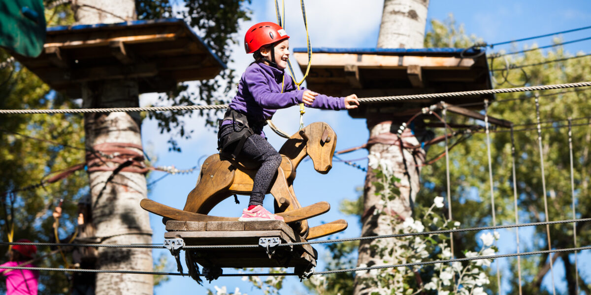 Ein Kind, das einen Helm und einen Klettergurt trägt, reitet auf einem hölzernen Schaukelpferd, das in einem Hochseilgarten in einem Outdoor-Abenteuerpark hängt.