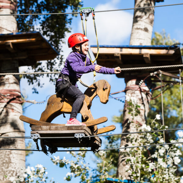 Ein Kind, das einen Helm und einen Klettergurt trägt, reitet auf einem hölzernen Schaukelpferd, das in einem Hochseilgarten in einem Outdoor-Abenteuerpark hängt.