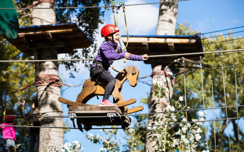 Ein Kind, das einen Helm und einen Klettergurt trägt, reitet auf einem hölzernen Schaukelpferd, das in einem Hochseilgarten in einem Outdoor-Abenteuerpark hängt.