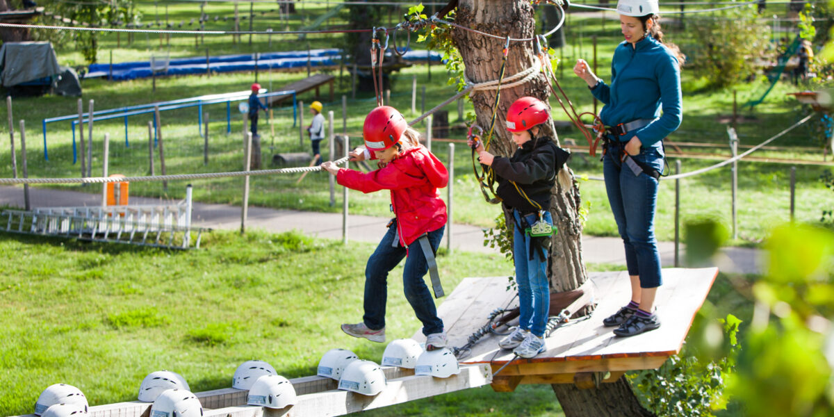 Zwei Kinder mit Helmen und Klettergurten überqueren unter Aufsicht eines Erwachsenen eine Holzplattform in einem Hochseilgarten. Bäume und Seile sind in der Umgebung des Parks zu sehen.