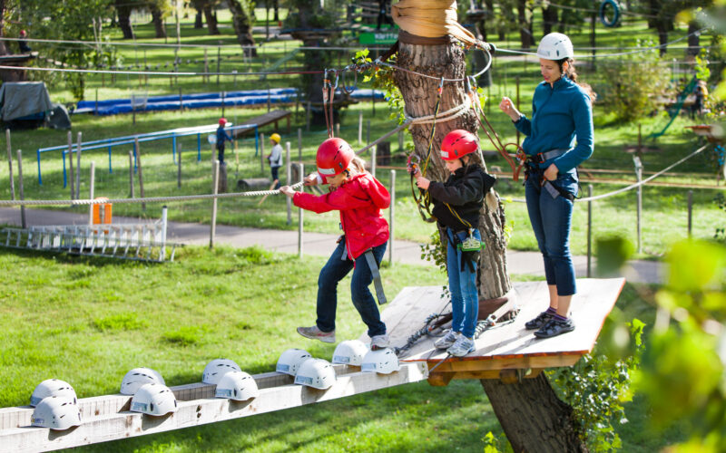Zwei Kinder mit Helmen und Klettergurten überqueren unter Aufsicht eines Erwachsenen eine Holzplattform in einem Hochseilgarten. Bäume und Seile sind in der Umgebung des Parks zu sehen.