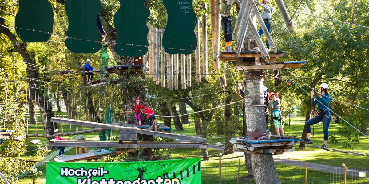 In einem Hochseilgarten in einem Park werden Seilbrücken und Hindernisse mit Klettergurten und Helmen überwunden. Auf einem grünen Schild steht "Hochseil-Klettergarten".