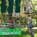 In einem Hochseilgarten in einem Park werden Seilbrücken und Hindernisse mit Klettergurten und Helmen überwunden. Auf einem grünen Schild steht "Hochseil-Klettergarten".