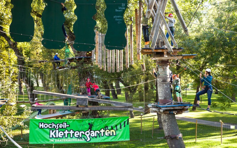 In einem Hochseilgarten in einem Park werden Seilbrücken und Hindernisse mit Klettergurten und Helmen überwunden. Auf einem grünen Schild steht "Hochseil-Klettergarten".