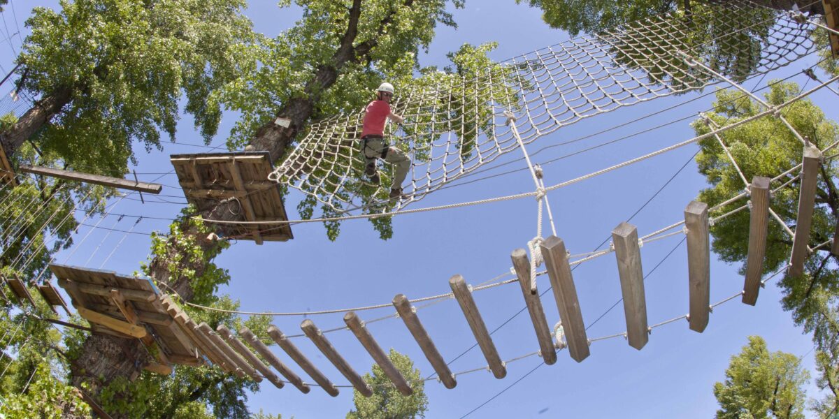 Eine Person, die einen Helm und einen Klettergurt trägt, überquert an einem sonnigen Tag ein Seilnetz hoch in den Bäumen eines Outdoor-Abenteuerparks.