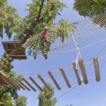 Eine Person, die einen Helm und einen Klettergurt trägt, überquert an einem sonnigen Tag ein Seilnetz hoch in den Bäumen eines Outdoor-Abenteuerparks.