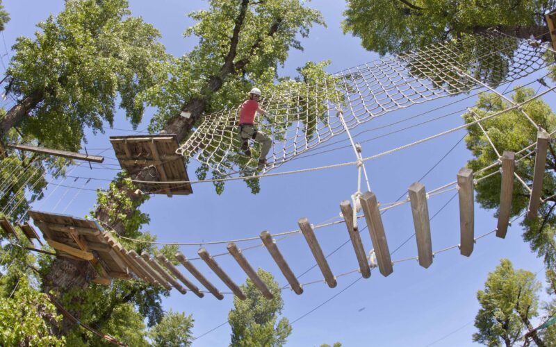 Eine Person, die einen Helm und einen Klettergurt trägt, überquert an einem sonnigen Tag ein Seilnetz hoch in den Bäumen eines Outdoor-Abenteuerparks.