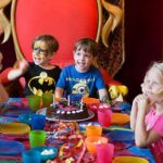 Eine Gruppe kleiner Kinder sitzt um einen geschmückten Tisch mit einem Schokoladengeburtstagskuchen, bunten Tassen und Partydekorationen. Zwei Jungen in der Mitte wirken aufgeregt und lächeln.