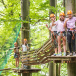 Vier Personen mit Helmen und Klettergurten stehen auf einer hölzernen Plattform in einem Waldabenteuerpark, während zwei andere auf einer entfernten Plattform warten, umgeben von grünen Bäumen.