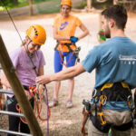 Eine Gruppe von Personen, die Helme und Klettergurte tragen, erhält von einem Ausbilder in einem Outdoor-Seilgarten Sicherheitsanweisungen.
