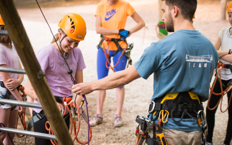 Eine Gruppe von Personen, die Helme und Klettergurte tragen, erhält von einem Ausbilder in einem Outdoor-Seilgarten Sicherheitsanweisungen.