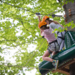 Zwei Jugendliche mit Helmen und Klettergurten sind an einer Seilrutsche in einem Wald befestigt und bereiten sich auf einen Abenteuerparcours in den Bäumen vor.