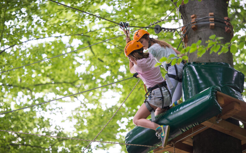 Zwei Jugendliche mit Helmen und Klettergurten sind an einer Seilrutsche in einem Wald befestigt und bereiten sich auf einen Abenteuerparcours in den Bäumen vor.