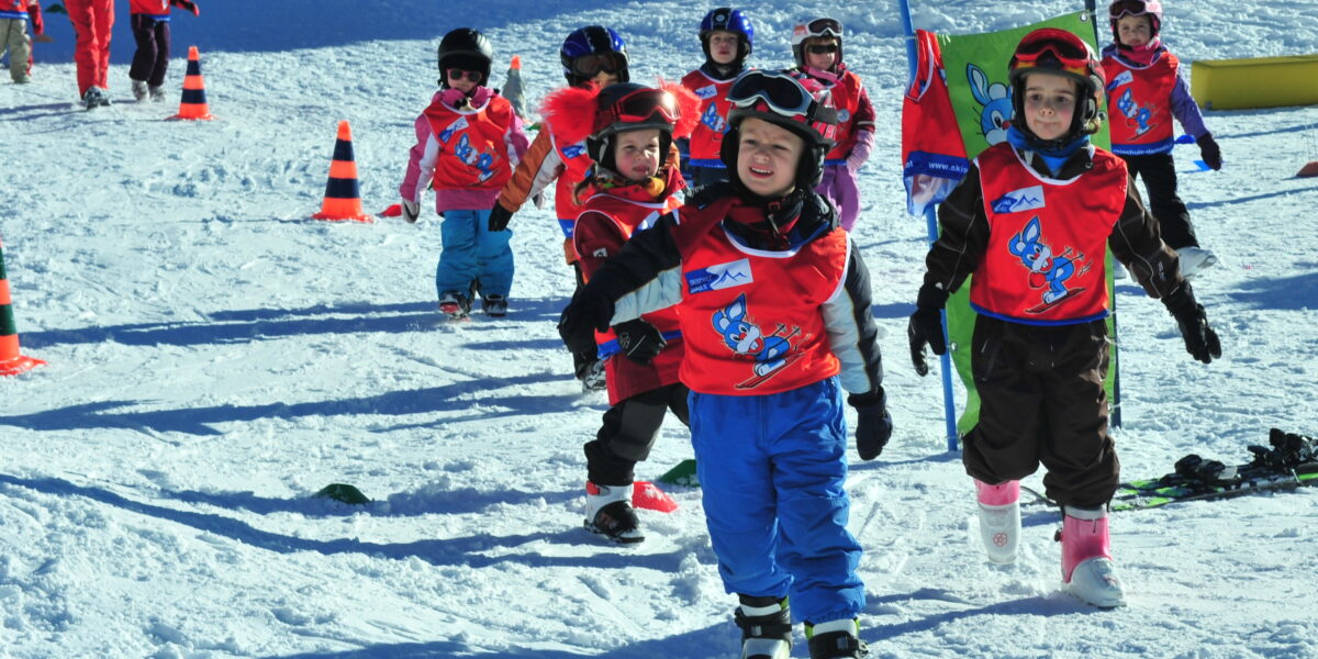 Eine Gruppe von Kindern in Skikleidung und roten Westen läuft in einer Skischule auf dem Schnee, während Lehrer und Sicherheitskegel in der Nähe sind.
