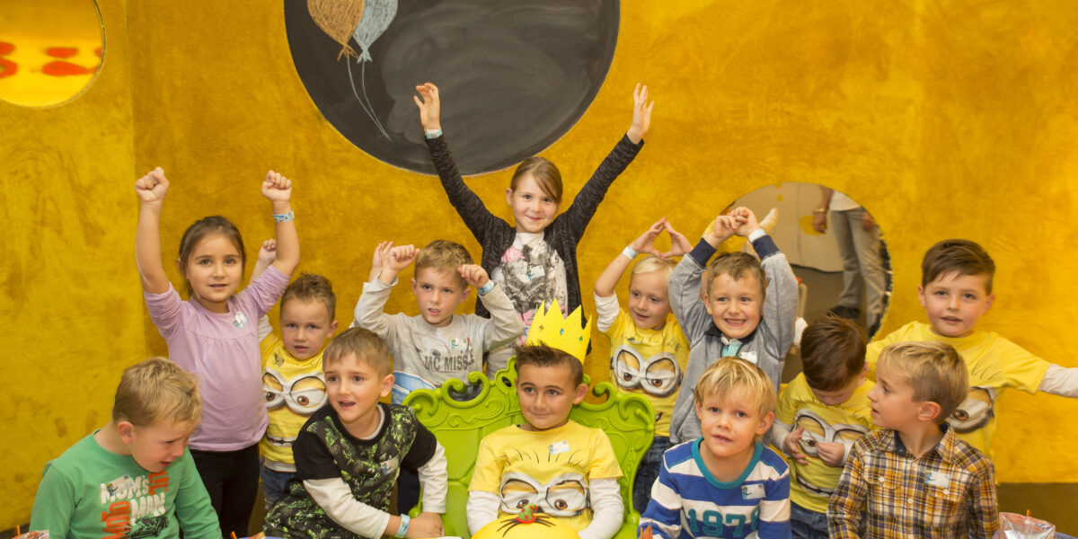 Eine Gruppe von Kindern auf einer Geburtstagsparty sitzt und steht um einen Tisch herum, der mit Minion-Motiven und einer Torte dekoriert ist, während an der Wand hinter ihnen eine "Happy Birthday"-Nachricht steht.