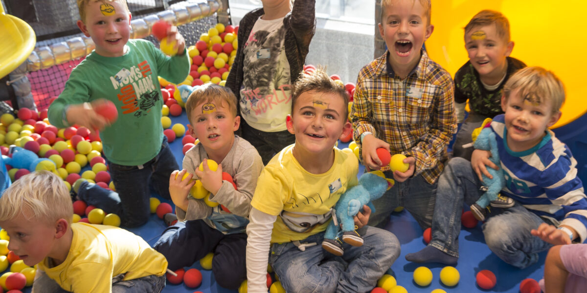 Eine Gruppe von Kindern mit Gesichtsbemalung spielt und lächelt in einer mit roten und gelben Plastikbällen gefüllten Ballgrube auf einem Indoor-Spielplatz.