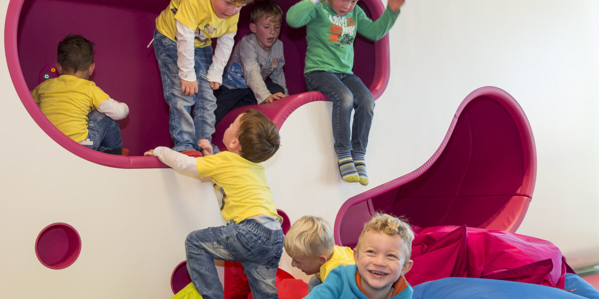Sechs kleine Kinder spielen auf bunten Sitzsäcken und klettern in ein großes, weißes Indoor-Spielgerät mit rosa Nischen hinein und wieder heraus.