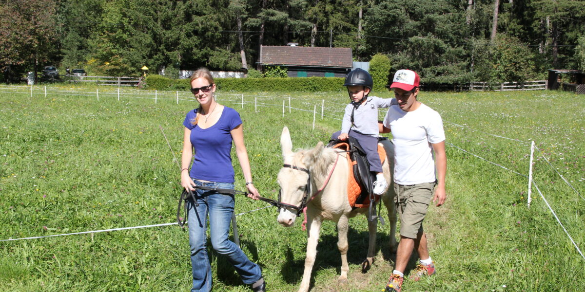 Ein Kind, das einen Helm trägt, reitet auf einem kleinen weißen Pferd, das von zwei Erwachsenen geführt wird, über einen grasbewachsenen Weg in einer ländlichen, bewaldeten Gegend.