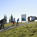 Drei Personen gehen auf einem Kiesweg bergauf in Richtung eines Informationsschildes und einer großen dekorativen Kofferkonstruktion auf einer Wiese mit Bäumen im Hintergrund.