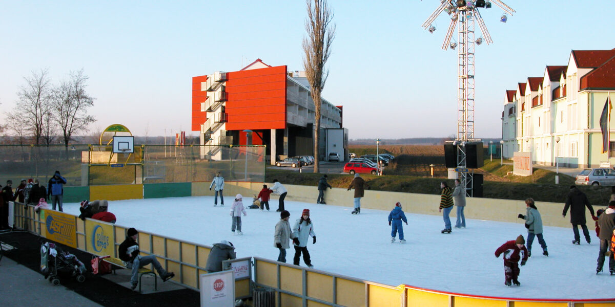 Menschen verschiedenen Alters, die auf einer Eisbahn im Freien Schlittschuh laufen, mit ein paar Schaulustigen in der Nähe; im Hintergrund sind Gebäude und eine große Beleuchtungsanlage zu sehen.