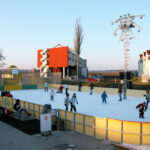 Menschen verschiedenen Alters, die auf einer Eisbahn im Freien Schlittschuh laufen, mit ein paar Schaulustigen in der Nähe; im Hintergrund sind Gebäude und eine große Beleuchtungsanlage zu sehen.