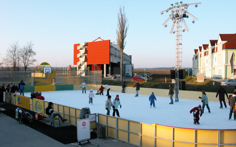 Menschen verschiedenen Alters, die auf einer Eisbahn im Freien Schlittschuh laufen, mit ein paar Schaulustigen in der Nähe; im Hintergrund sind Gebäude und eine große Beleuchtungsanlage zu sehen.