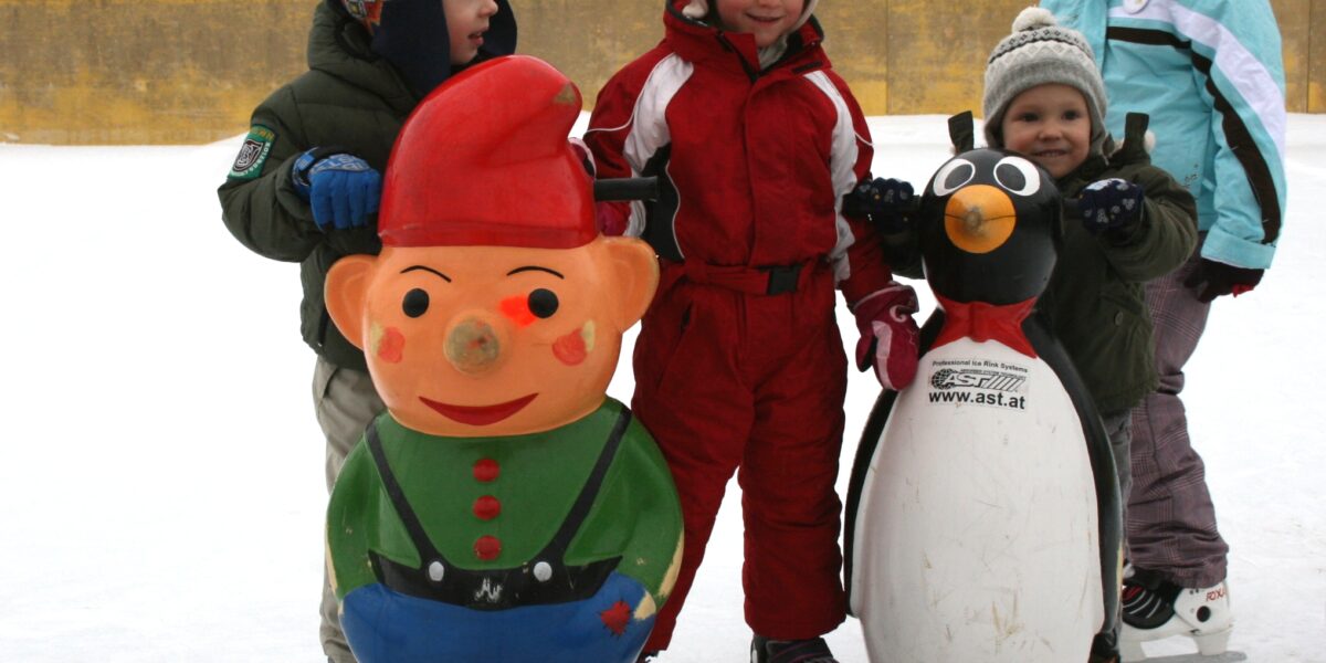 Vier Kinder, die im Freien Schlittschuh laufen, benutzen vor einem Hotelgebäude Eislaufhilfen in Form von Zeichentrickfiguren, darunter ein Zwerg und ein Pinguin.