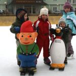 Vier Kinder, die im Freien Schlittschuh laufen, benutzen vor einem Hotelgebäude Eislaufhilfen in Form von Zeichentrickfiguren, darunter ein Zwerg und ein Pinguin.
