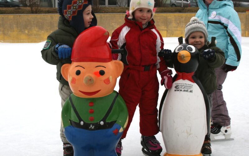 Vier Kinder, die im Freien Schlittschuh laufen, benutzen vor einem Hotelgebäude Eislaufhilfen in Form von Zeichentrickfiguren, darunter ein Zwerg und ein Pinguin.