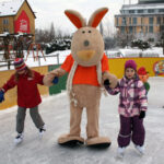 Zwei Kinder halten sich auf einer Eisbahn im Freien mit einer Person im Kängurukostüm an den Händen. Im Hintergrund sind eine Pinguin-Eislaufhilfe und Spielgeräte zu sehen.