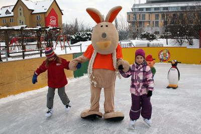 Zwei Kinder halten sich auf einer Eisbahn im Freien mit einer Person im Kängurukostüm an den Händen. Im Hintergrund sind eine Pinguin-Eislaufhilfe und Spielgeräte zu sehen.