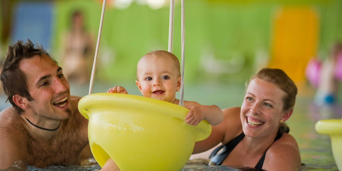 Ein Baby sitzt lächelnd in einer gelben Schwimmhilfe in einem Schwimmbecken, daneben ein erwachsener Mann und eine erwachsene Frau im Wasser.