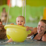Ein Baby sitzt lächelnd in einer gelben Schwimmhilfe in einem Schwimmbecken, daneben ein erwachsener Mann und eine erwachsene Frau im Wasser.