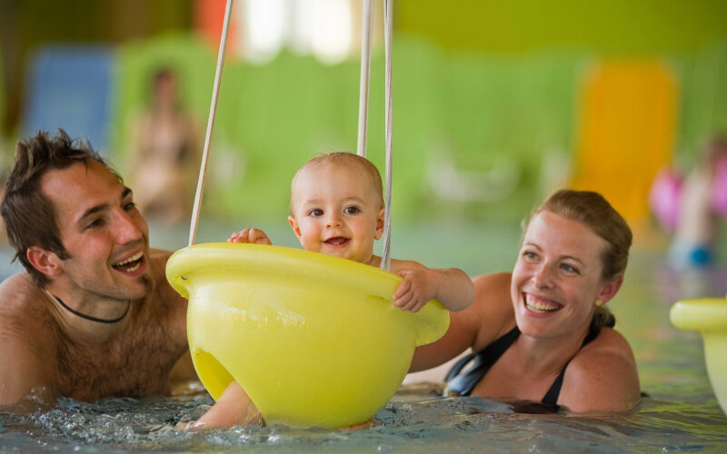 Ein Baby sitzt lächelnd in einer gelben Schwimmhilfe in einem Schwimmbecken, daneben ein erwachsener Mann und eine erwachsene Frau im Wasser.
