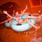 Zwei Kinder in Badekleidung fahren mit blauen aufblasbaren Schläuchen eine Indoor-Wasserrutsche hinunter und lächeln mit erhobenen Armen, während das Wasser um sie herum spritzt.