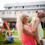 Eine Frau hält ein lachendes kleines Mädchen im Freien in der Nähe eines Spielplatzes, mit einem modernen Gebäude und Menschen, die im Gras sitzen, im Hintergrund.