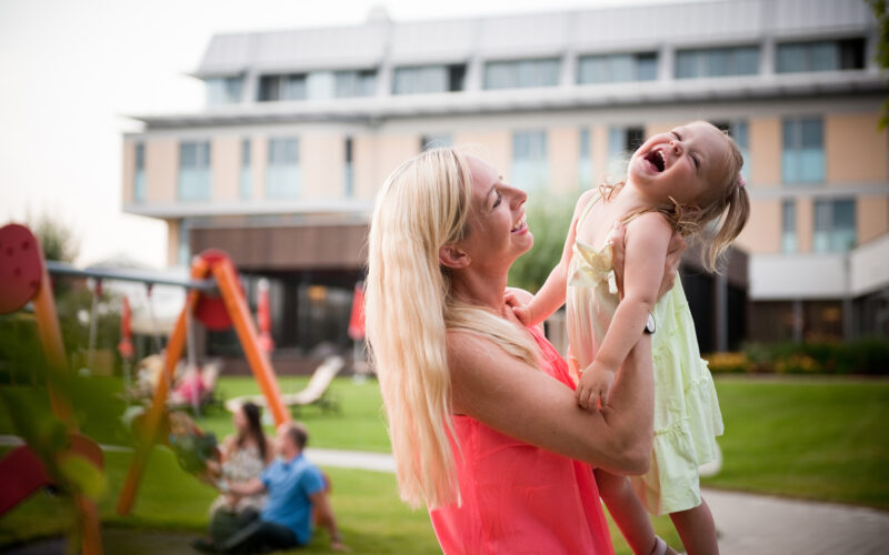 Eine Frau hält ein lachendes kleines Mädchen im Freien in der Nähe eines Spielplatzes, mit einem modernen Gebäude und Menschen, die im Gras sitzen, im Hintergrund.
