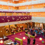 Großes, farbenfrohes Foyer mit Gruppen von Menschen, meist Kindern, die unter aufgehängten dekorativen Kugeln versammelt sind. Moderne Sitzmöbel und eine Wanduhr sind zu sehen. Der Raum ist hell beleuchtet.