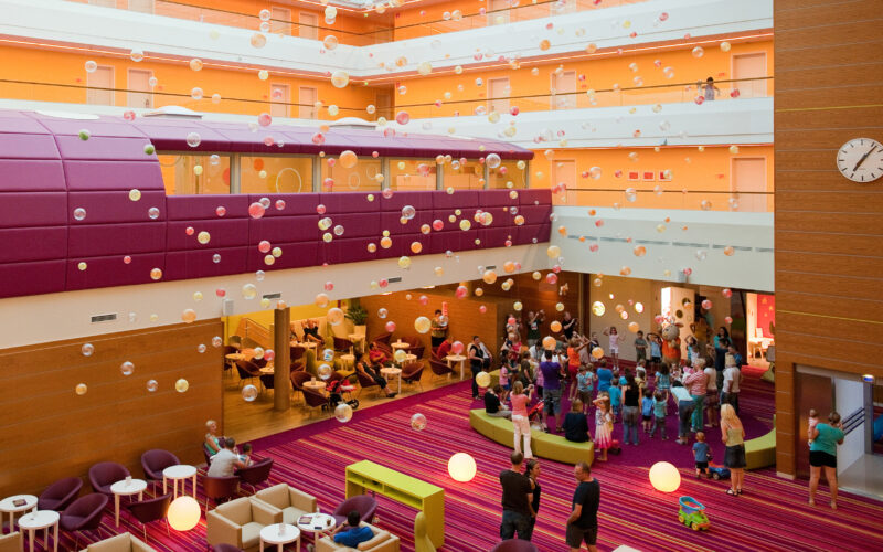 Großes, farbenfrohes Foyer mit Gruppen von Menschen, meist Kindern, die unter aufgehängten dekorativen Kugeln versammelt sind. Moderne Sitzmöbel und eine Wanduhr sind zu sehen. Der Raum ist hell beleuchtet.