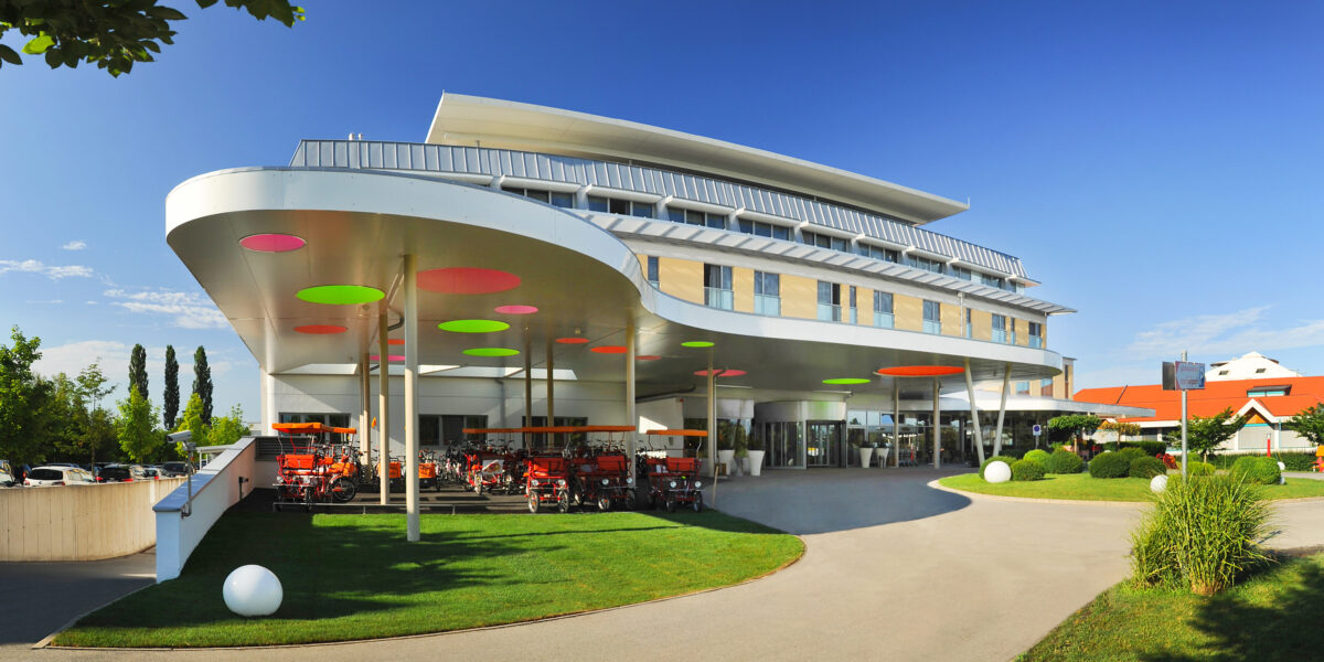 Modernes Gebäude mit geschwungener Architektur, bunten, kreisförmigen Lichtern an der Decke, Rasen und mehreren roten Kinderwagen, die vor dem Eingang geparkt sind, unter einem sonnigen Himmel.
