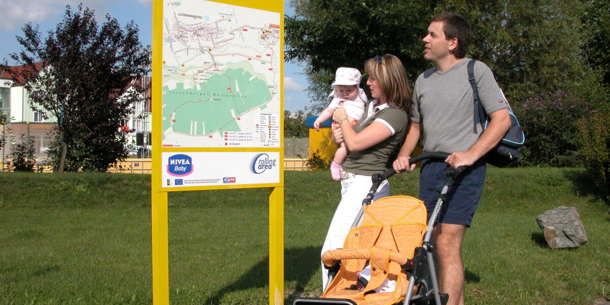 Eine Familie mit Kinderwagen und Baby steht an einem sonnigen Tag in einer Parkanlage neben einem großen gelben Schild mit der Aufschrift „Baby on Tour“.