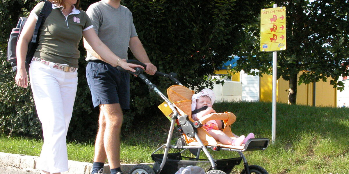 Eine Frau, ein Mann und ein Baby im Kinderwagen gehen an einem sonnigen Tag auf einem gepflasterten Weg in der Nähe eines gelben Schildes und Bäumen spazieren.